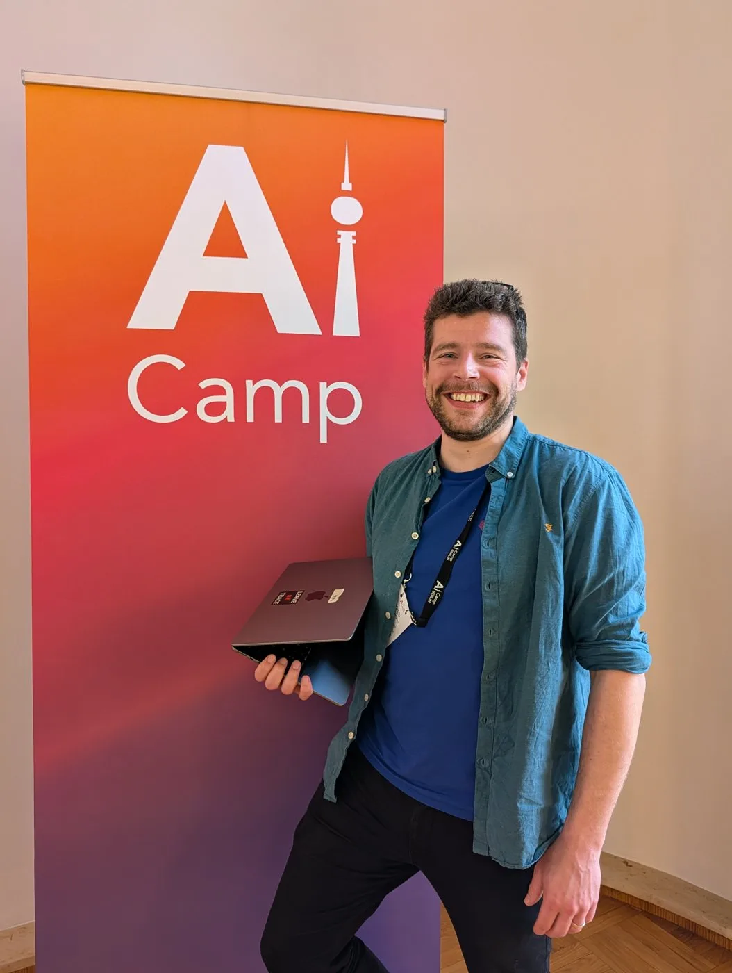 Waldo smiling in front of the orange-to-purple AI Camp Berlin roll-up banner, holding a laptop and wearing an AI Camp lanyard