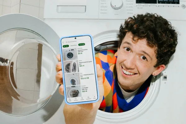 A smiling person looks out of a washing machine drum and holds up a phone showing Kleinanzeigen search results for used washing machines