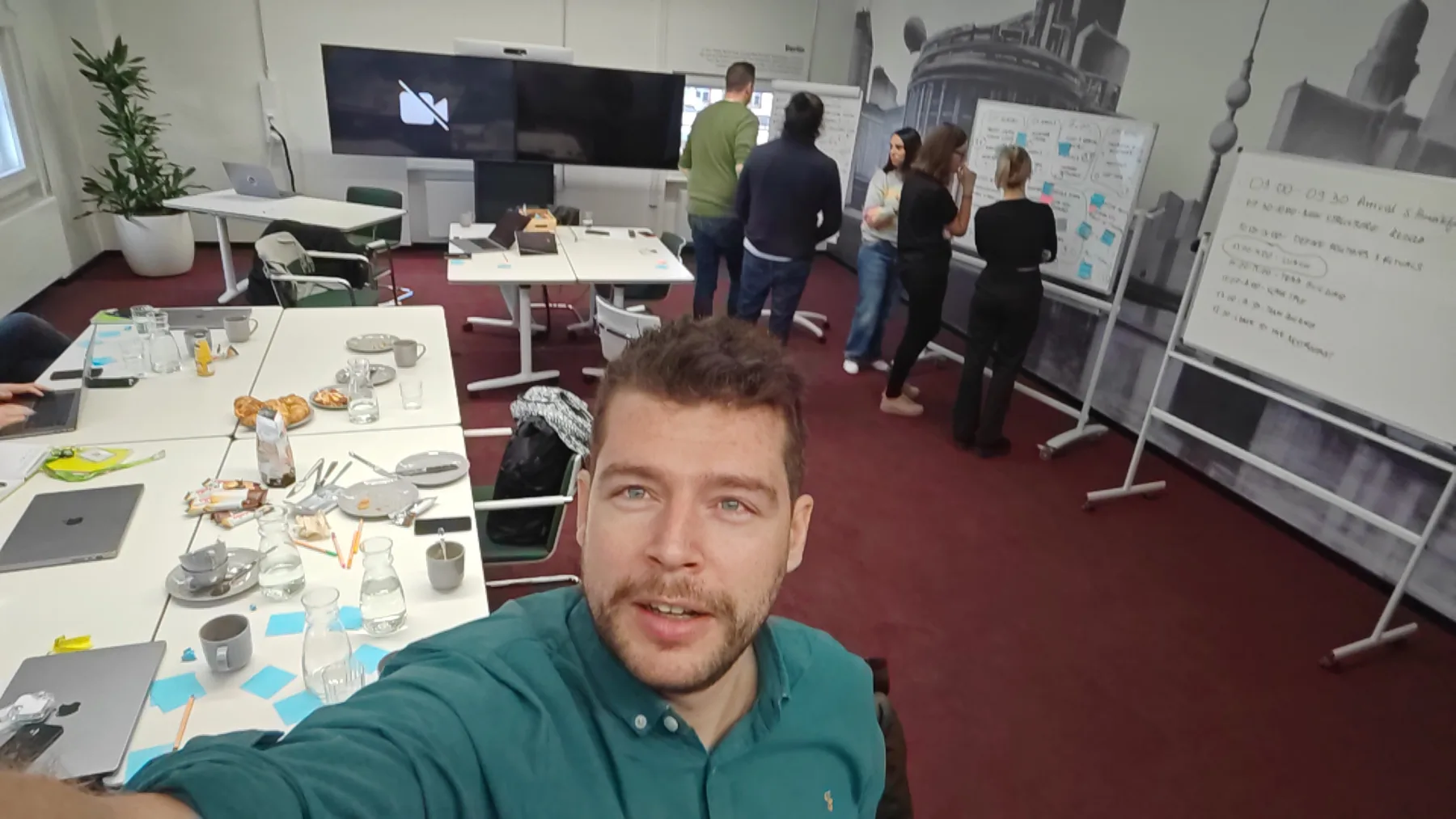 A workshop room with a team standing at two whiteboards covered in sticky notes, coffee cups and croissants on the tables, and the author taking a selfie in the foreground