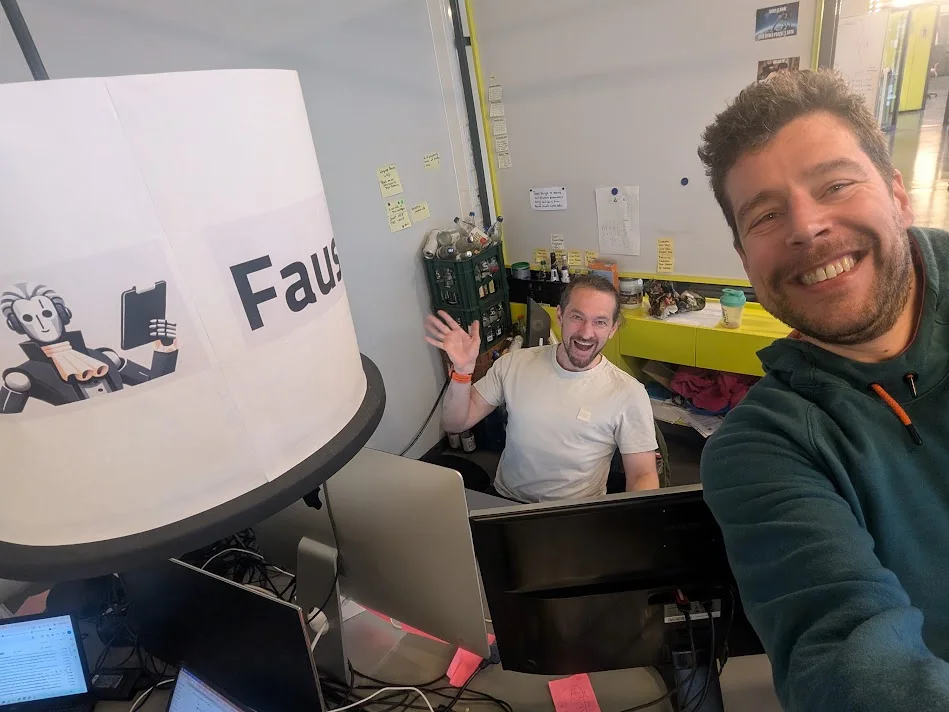 Waldo and a Faust AI colleague at their desks, a printed Faust AI sign in the foreground
